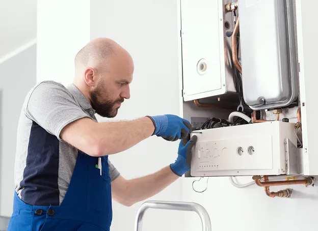 Technicien en salopette bleue et gants, répare une chaudière murale avec un tournevis.